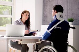 Man man sitting in a wheelchair and wearing a neckbrace in front of a desk, and the woman explaining if the man can quit their job while on workers’ compensation