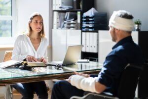 Man with a bandage wrapped around his forehead sitting in an office in front of a desk speaking to a woman on the other side of the desk who is explaining how to check the status of a workers’ compensation claim