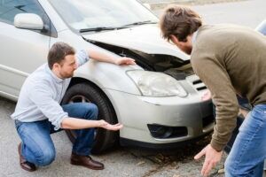 Two people standing next to the front of a car that was damaged in an auto accident, with both individuals wondering if they should call the police for a fender bender