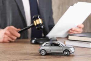 A lawyer sits at a desk near a gavel and a toy car, ready to explain why victims should get a lawyer for a car accident.