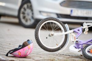A close-up shot of a child’s bicycle and helmet on the ground in front of a car. A nearby parent wonders what the average settlement for a child in a car accident is. 